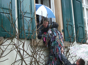 Person guckt mit Regenschirm aus einem Fenster, verkleideter Mensch redet mit der Person