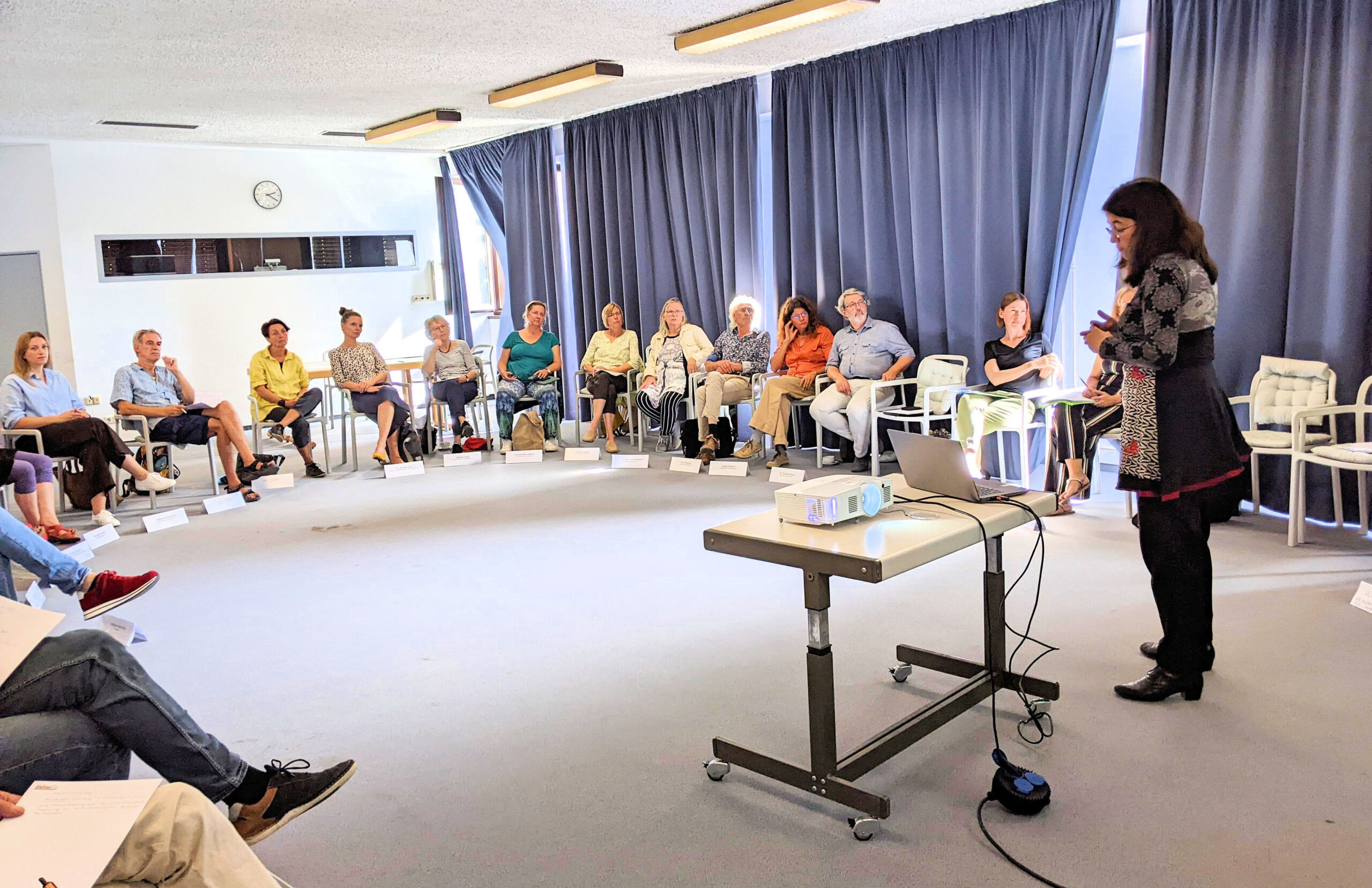 Stuhlkreis mit Teilnehmenden aus dem Arbeitskreis Soziokultur und Kulturelle Bildung. Im Vordergrund steht Ulrike Blumenreich und präsentiert mit Laptop und Beamer die Bausteine der Kulturentwicklungsplanung.