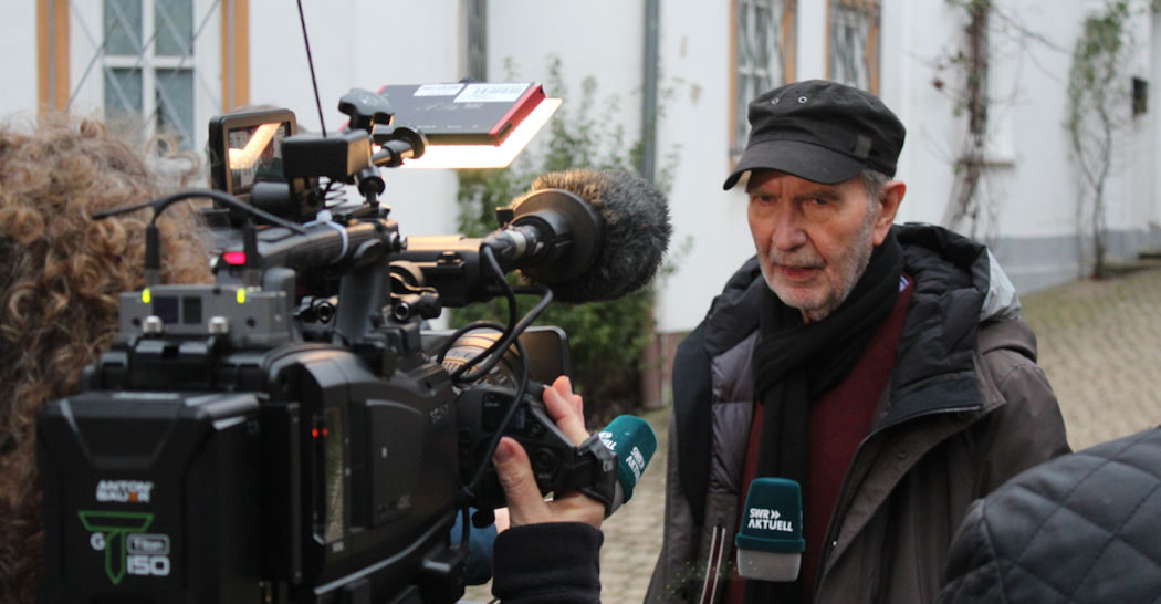 Der Regisseur Edgar Reitz steht vor einer Kamera und spricht in die Kamera.