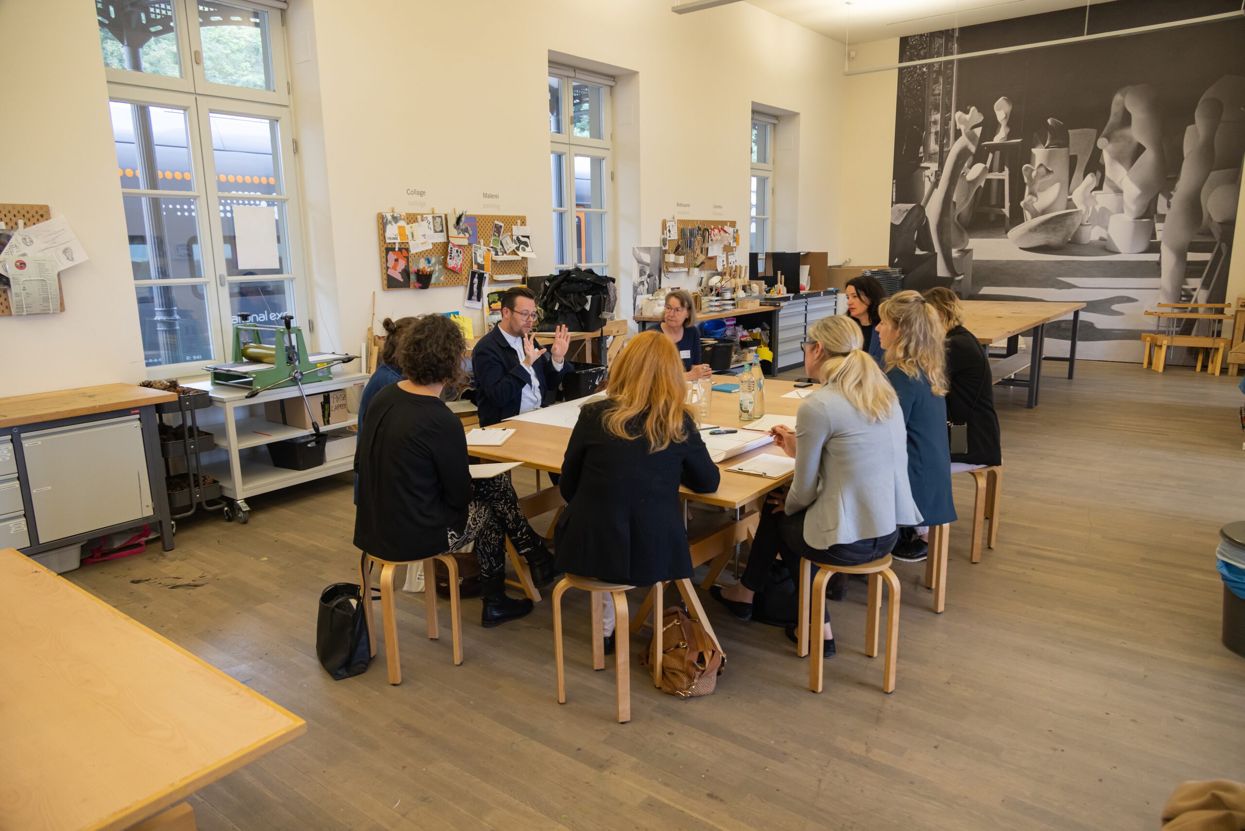 Blick auf eine Arbeitsgruppe. Die Teilnehmenden sitzen auf Hockern um einen Werkstatttisch