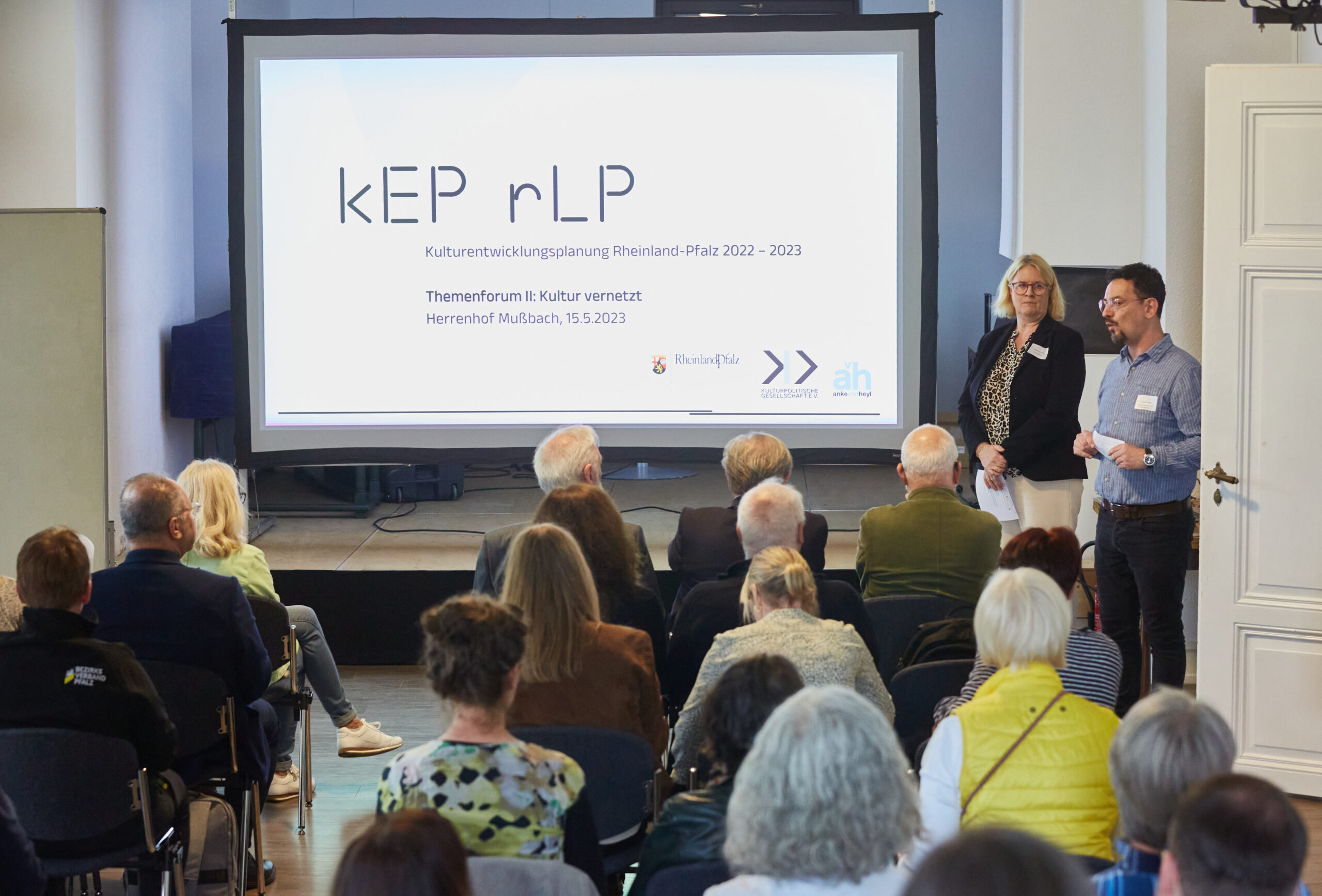 Auftakt zum zweiten Themenforum der Kulturentwicklungsplanung in Rheinland-Pfalz. Vorne rechts steht Roderick Haas vor den Teilnehmenden in Sitzreihen. Im Hintergrund eine Präsentation, auf der steht: KEP rlp. Kulturentwicklungsplanung Rheinland-Pfalz 2022-2023. Themenforum II: Kultur vernetzt. Herrenhof Mußbach