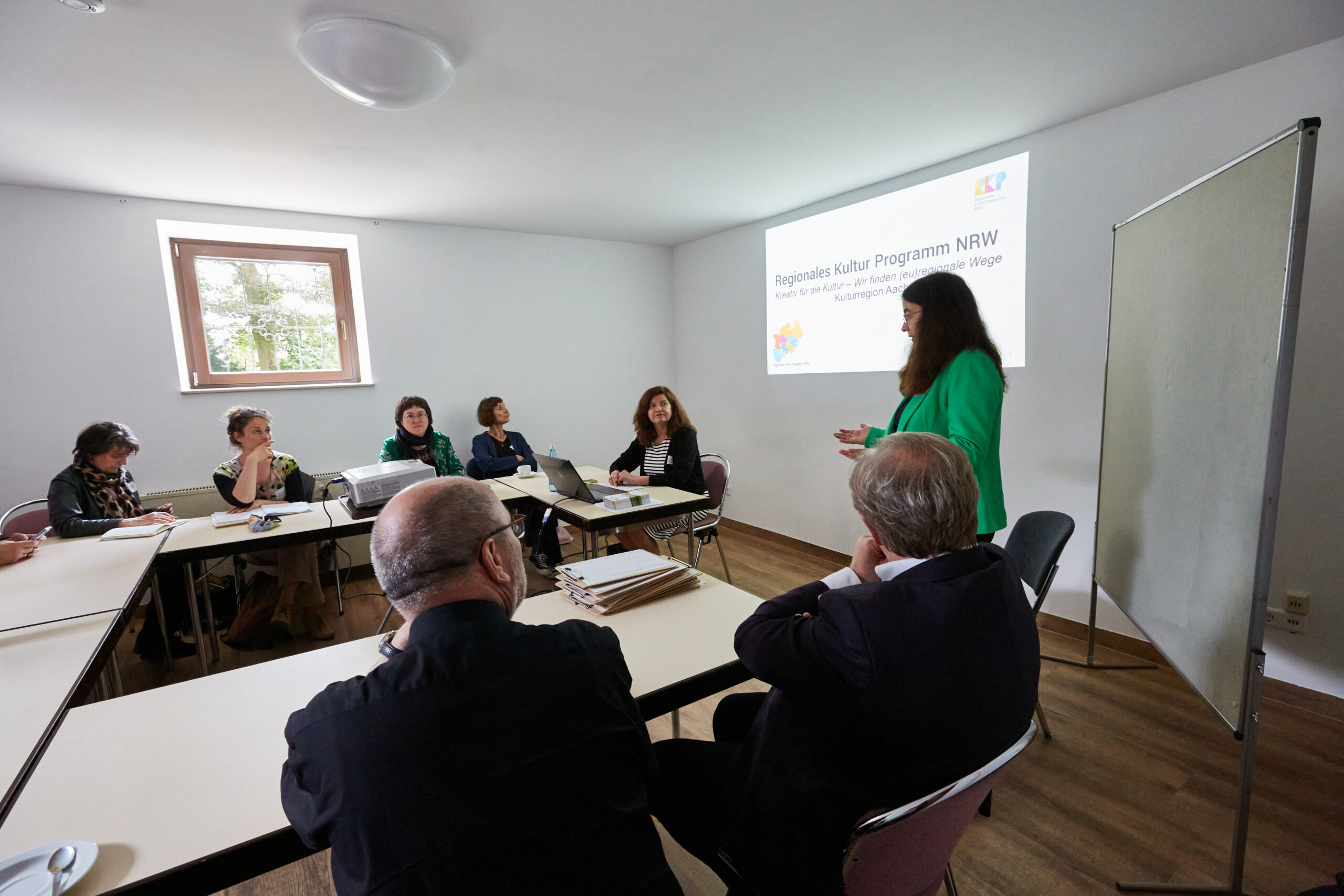 Diskussionsrunde beim zweiten Themenforum. An einem U aus Tischen sitzen (erkennbar) 8 Personen. Eine Person in grüner Jacke steht vor einer Metaplanwand und hält einen Vortrag. An der Wand sieht man eine Präsentation, auf der steht: Regionales Kultur Programm NRW