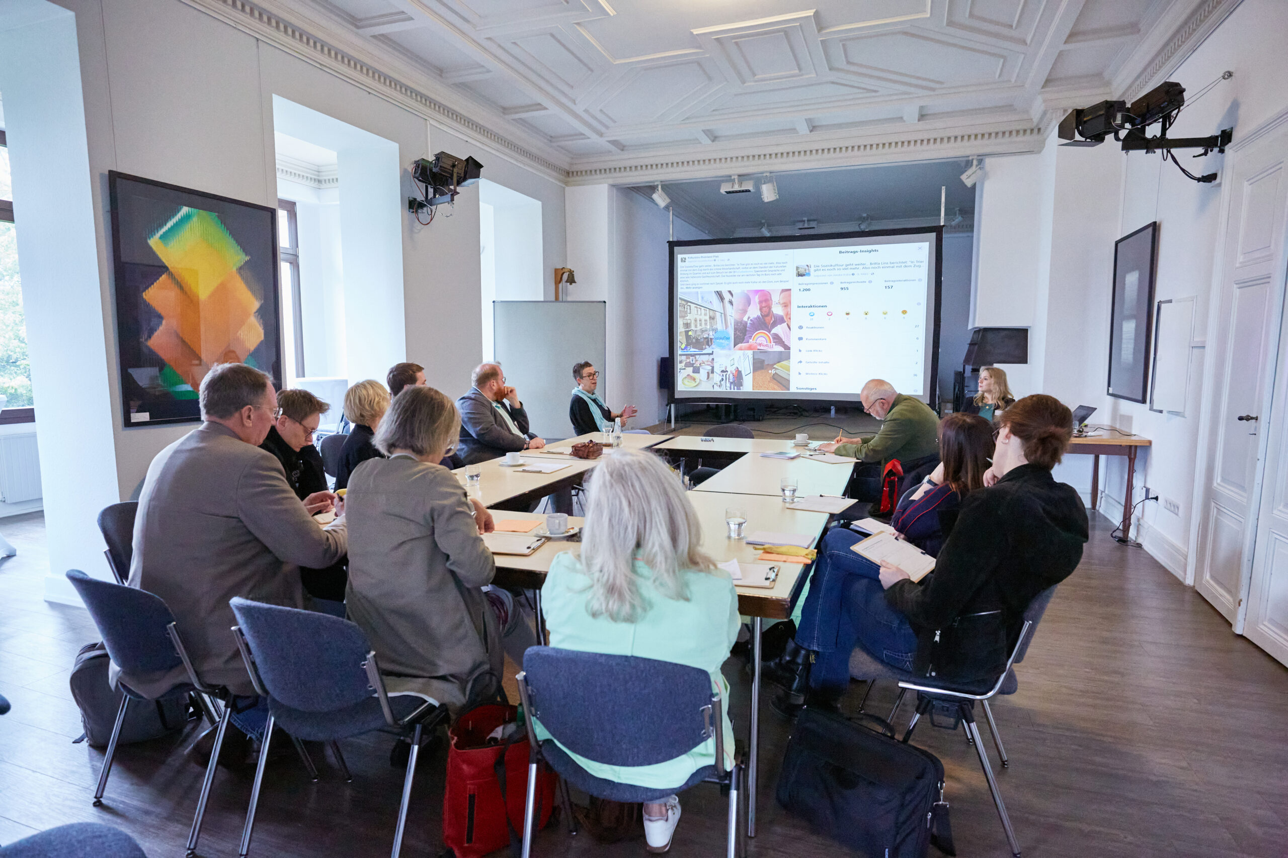 Blick auf eine Diskussionsrunde beim zweiten Themenforum der Kulturentwicklungsplanung. Ein Rechteck aus Tischen. Daran sitzen 12 Menschen und schauen auf eine Leinwand. Auf der läuft ein Präsentation.