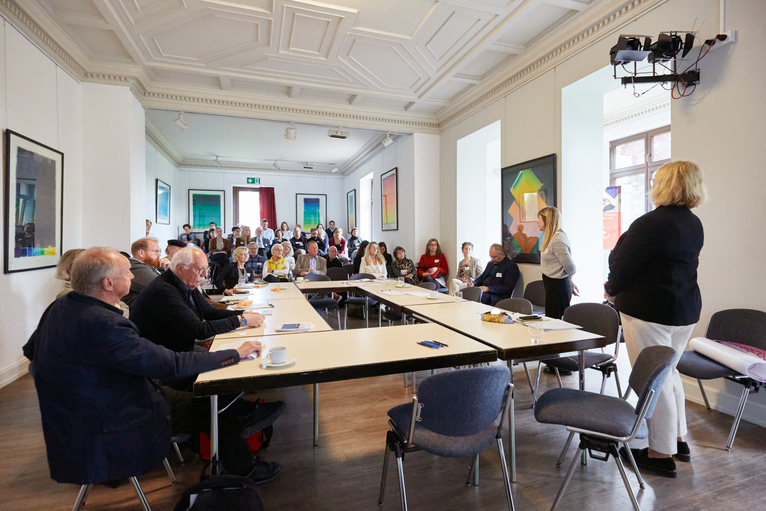 Finale des zweiten Themenforums der Kulturentwicklungsplanung. Hinten sitzen die Teilnehmenden in Stuhlreihen. Vorne stehen einige Tische zum Rechteck gestellt. An den Wänden hängt Kunst. Nach oben blickt man auf eine aufwändig gestaltete Holzdecke