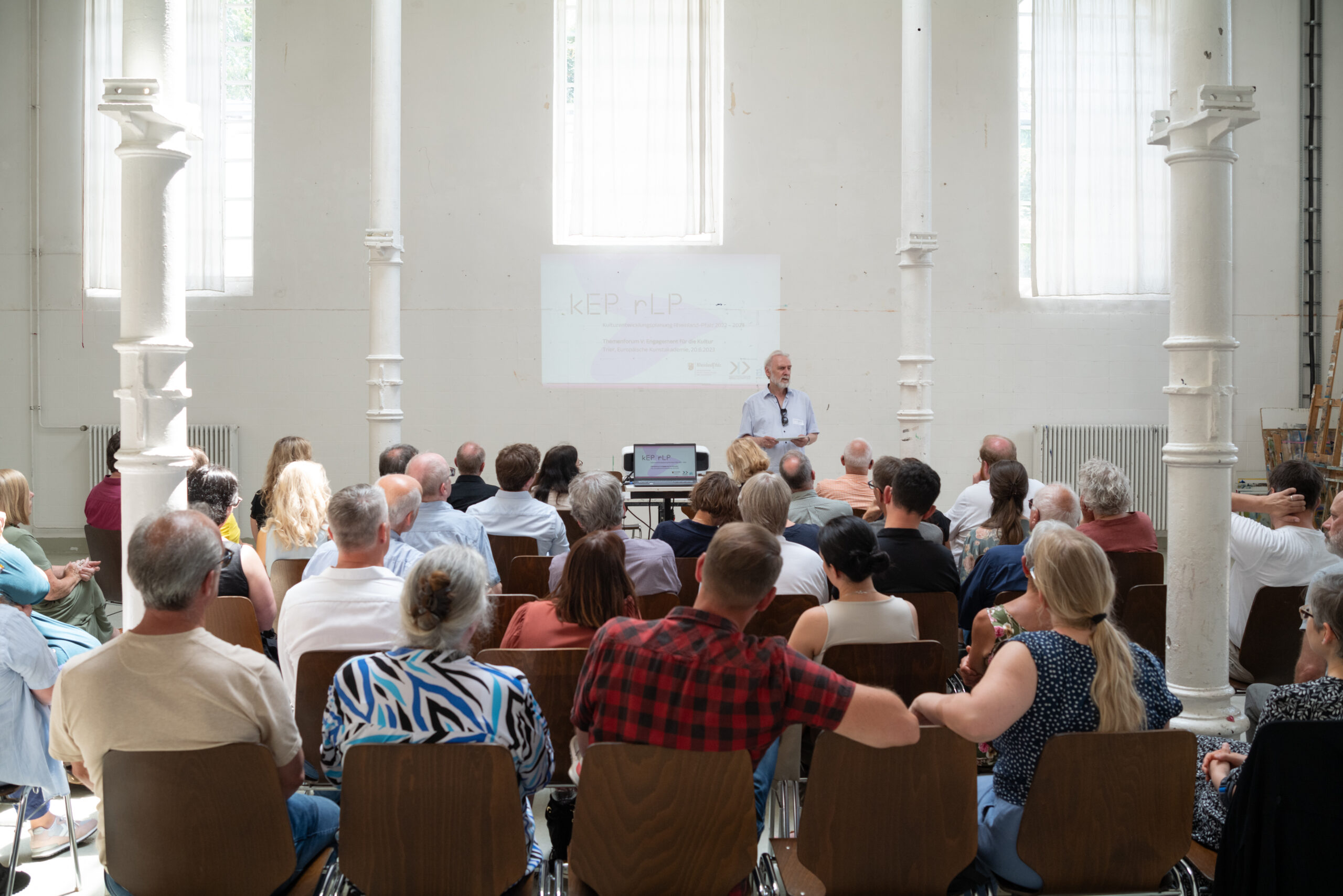 Große Gruppe in Sitzreihen in einer hellen Halle. Vorne steht ein Mann, der eine Begrüßungsrede vorträgt.