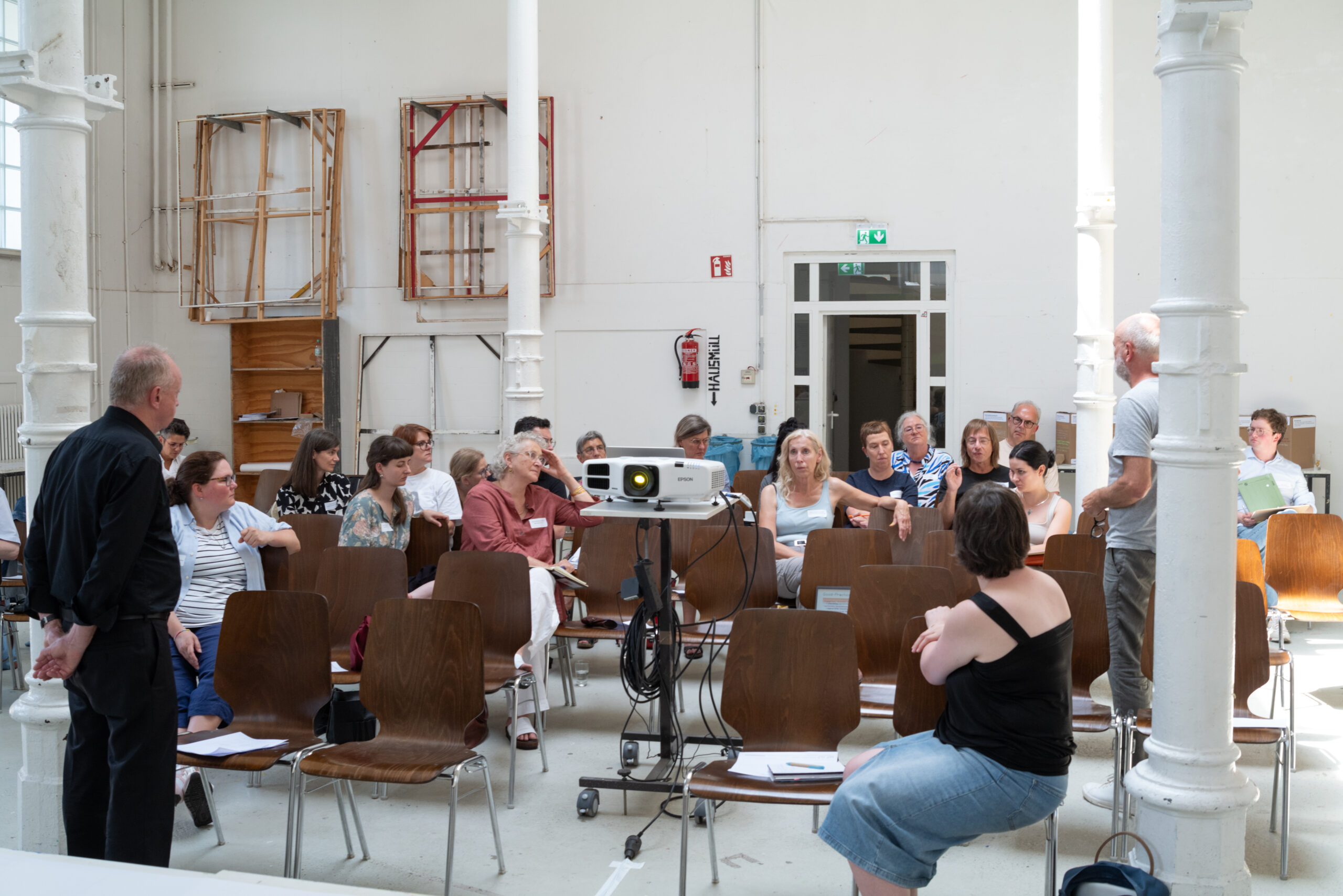 Blick in eine Gruppe miteinander diskutierender Menschen in einer hellen Halle. Man sieht einen Beamer. Die Halle wird von Metallsäulen gestürzt. An der Wand hängen Keilrahmen