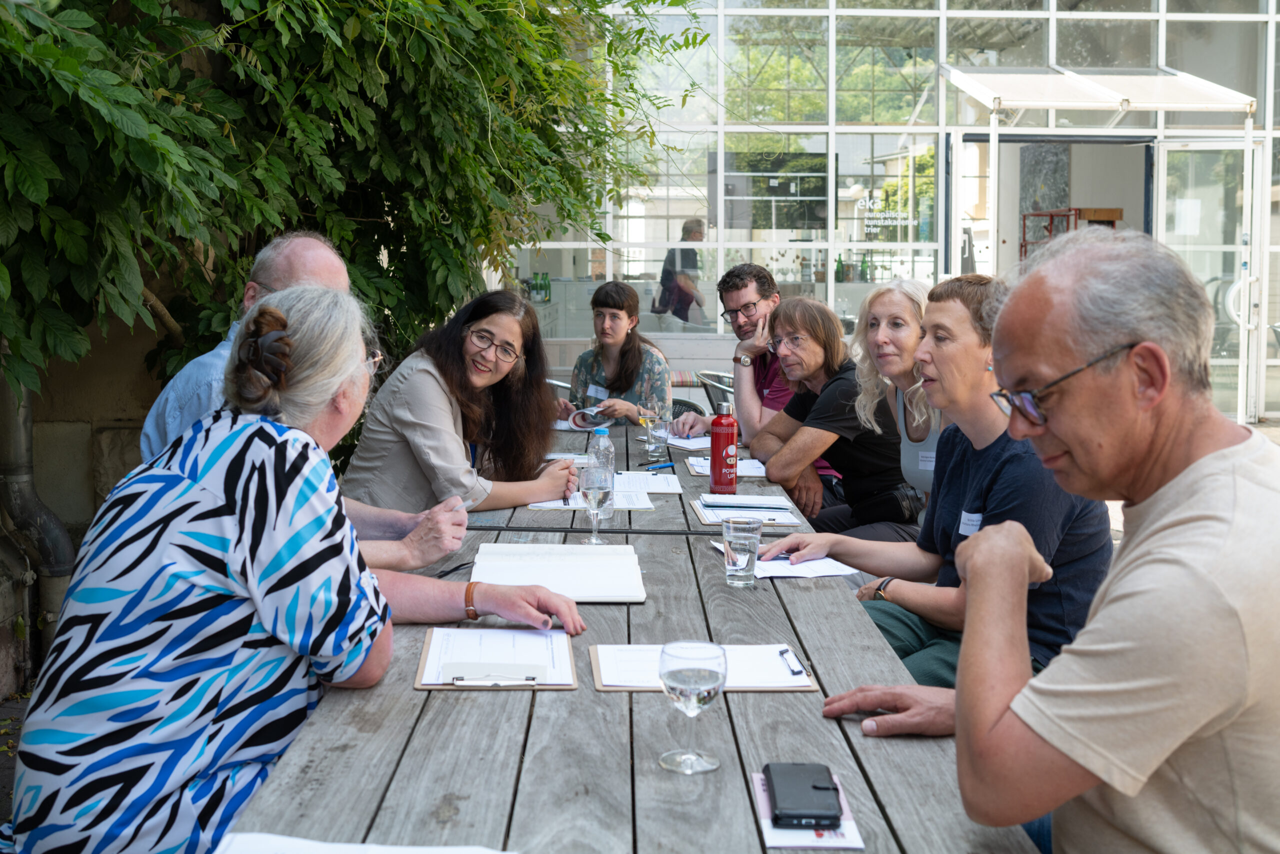 Diskussion von einer Gruppe an einem Holztisch, die im Freien sitzen und Papier und Stifte vor sich liegen haben.